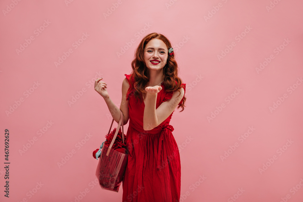Slim cute caucasian young woman in red summer dress smiling with teeth on pink background. Beautiful woman with long curly hair stretches her hand forward with bag in hands. Slim cute caucasian young woman in red summer dress smiling with teeth on pink background. Beautiful woman with long curly hair stretches her hand forward with bag in hands.