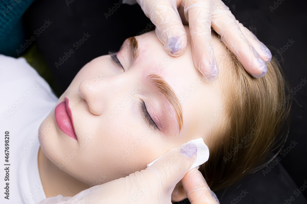 the girl model at the eyebrow lamination procedure, the master erases the remnants of their laminating compositions with a cotton sponge the girl model at the eyebrow lamination procedure, the master erases the remnants of their laminating compositions with a cotton sponge
