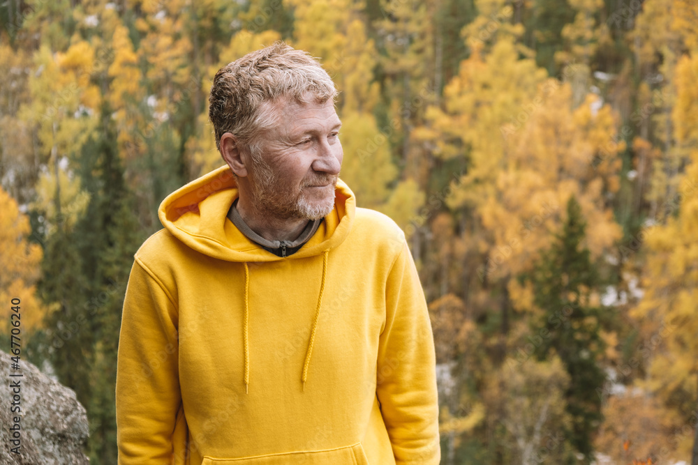 An authentic portrait of a smiling, unshaven middle-aged traveler in a yellow hoodie on top of a mountain against the background of an autumn forest. Close-up. Selective focus. Sustainable environment