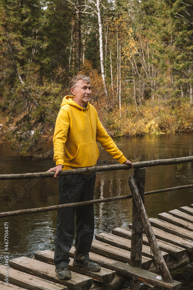 Local travel. A middle-aged traveler in yellow hoodie stands on wooden bridge over a river in the forest, enjoying beautiful autumn day outdoors. Sustainable environment. Eco tourism.