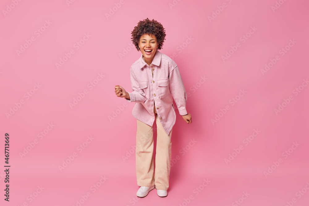 Full length shot of happy carefree woman with curly hair dances and moves actively wears stylish jacket trousers and sneakers has fun isolated over pink background. People happiness and joy concept Full length shot of happy carefree woman with curly hair dances and moves actively wears stylish jacket trousers and sneakers has fun isolated over pink background. People happiness and joy concept