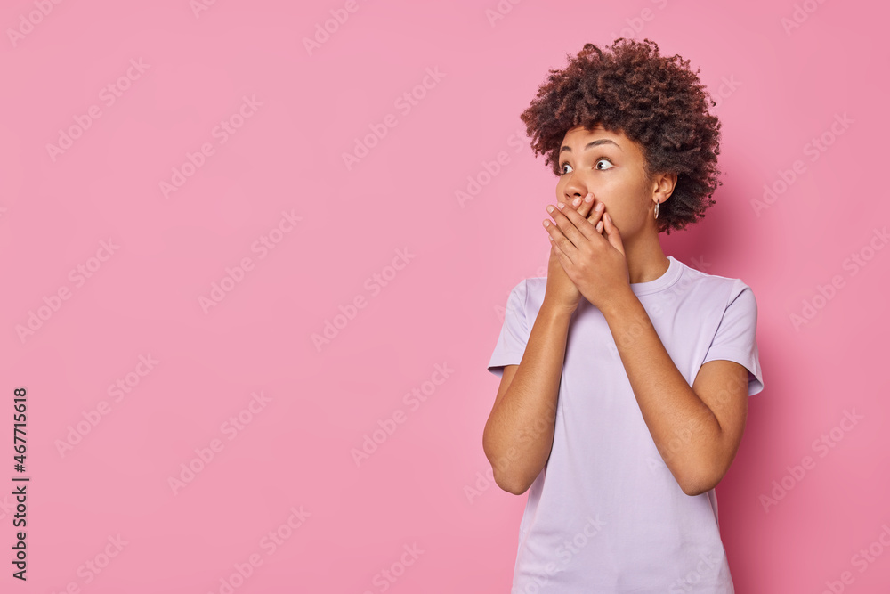 Photo of scared young curly woman covers mouth with hands looks terrified notices something horrible wears casual t shirt isolated over pink backgound copy space for your advertising content Photo of scared young curly woman covers mouth with hands looks terrified notices something horrible wears casual t shirt isolated over pink backgound copy space for your advertising content