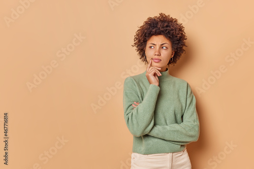 Serious curly haired woman stands in thoughtful pose concentrated away ponders on something important poses against beige background with blank space for your advertising content or promotion © Wayhome Studio
