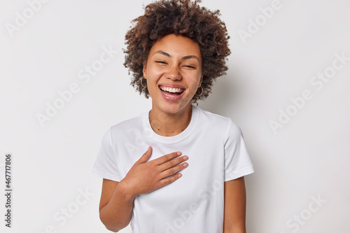 Portrait of happy sincere woman with curly hair keeps hand on chest expresses positive feelings hears heartwaring words dressed in casual t shirt isolaed over white background. Happiness concept