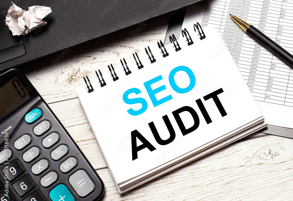 On a light-colored office table is a computer, a black calculator, a pen, and a notebook with the text SEO AUDIT. Business concept. On a light-colored office table is a computer, a black calculator, a pen, and a notebook with the text SEO AUDIT. Business concept.