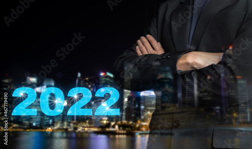 2022 new year business marketing concept. Double exposure businessman in black suit standing with his arms crossed and datum 2022 on blur night city background. Financial investment, growth planning