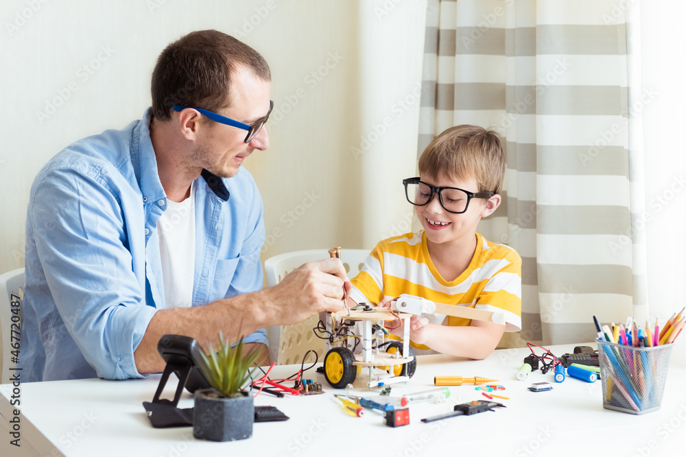 Father and son sitting by the white table and building robot at home as a school science project and having fun Father and son sitting by the white table and building robot at home as a school science project and having fun