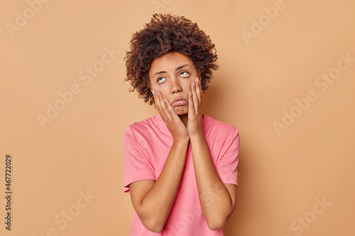 Fotografi Young curly haired woman has tired and bored look keeps palms on face fed up of