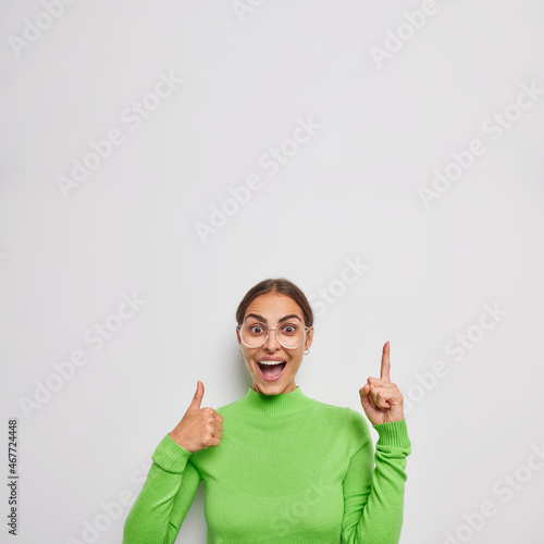 Vertical shot of happy brunette woman points above with index finger keeps thumb up praises excellent job recommends something dressed in casual green poloneck isolated over white background photo