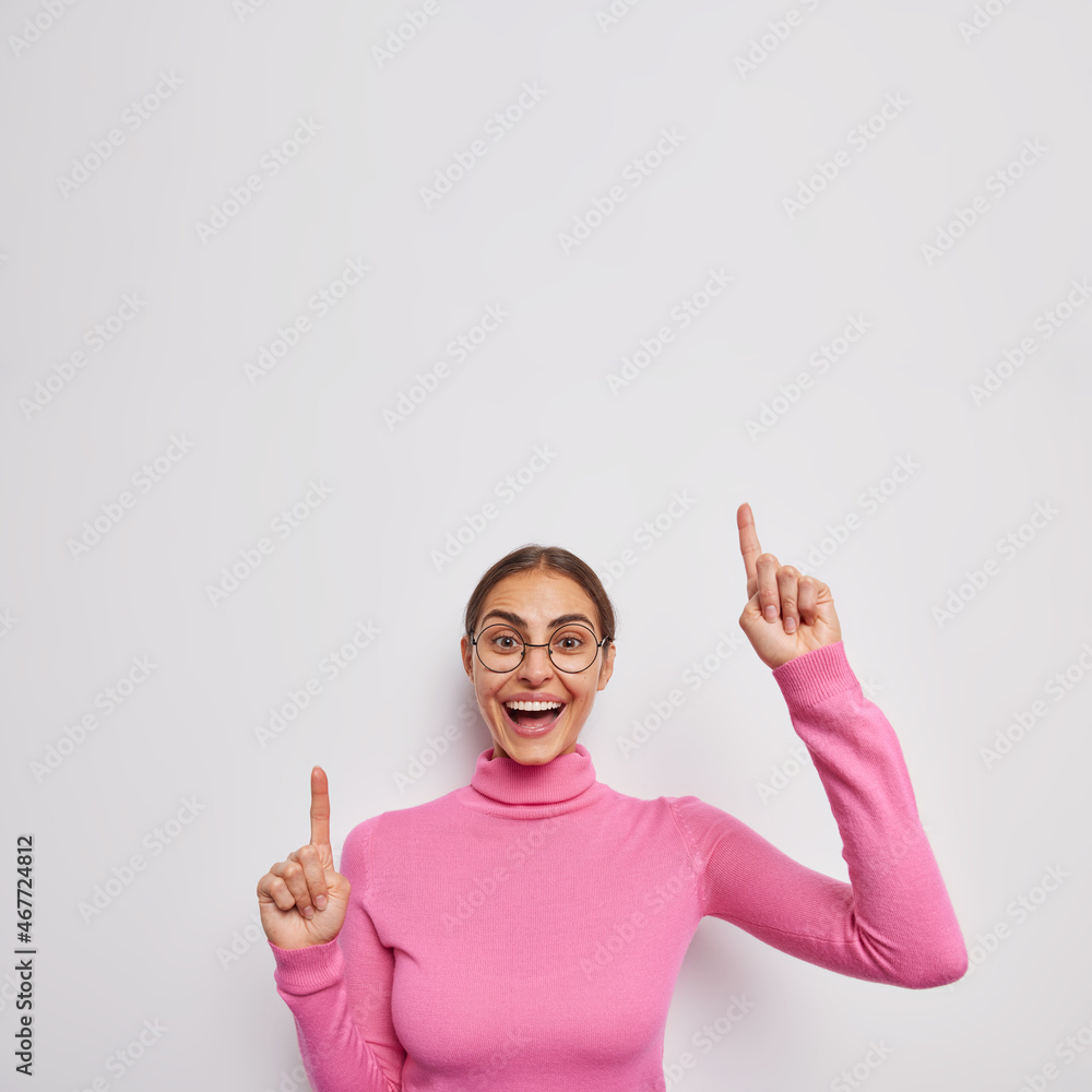 Positive young woman has playful mood points overhead shows awesome logo demonstrates advertisement looks happily isolated over white background. Happy positive female model promots something Positive young woman has playful mood points overhead shows awesome logo demonstrates advertisement looks happily isolated over white background. Happy positive female model promots something