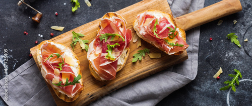 Open ham sandwiches, arugula and hard cheese, served on wooden stand with aglass of red wine on aconcrete old dark background. Rustik style. Top view