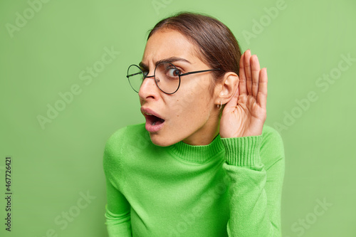 Indignant brunette woma tries to overhear someone holds hand near ear tries to understand words eavesdropping keeps mouth opened wears spectacles and poloneck isolated over green background. photo