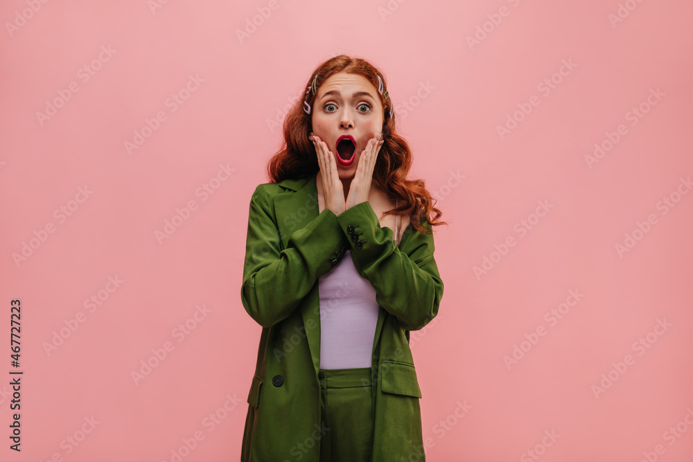 Pretty young woman yells in surprise with mouth wide open, holding cheeks with her hands. In closed room red-haired girl in lilac top and dark green suit. Reaction to unpleasant situation, concept Pretty young woman yells in surprise with mouth wide open, holding cheeks with her hands. In closed room red-haired girl in lilac top and dark green suit. Reaction to unpleasant situation, concept