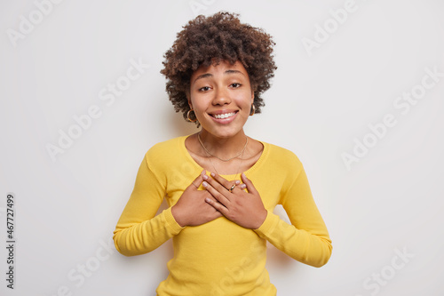Thankful smiling woman presses hands on chest has something in heart makes sweet memories gesture looks with appreciation wears yellow sweater poses against white background. Thank you very much