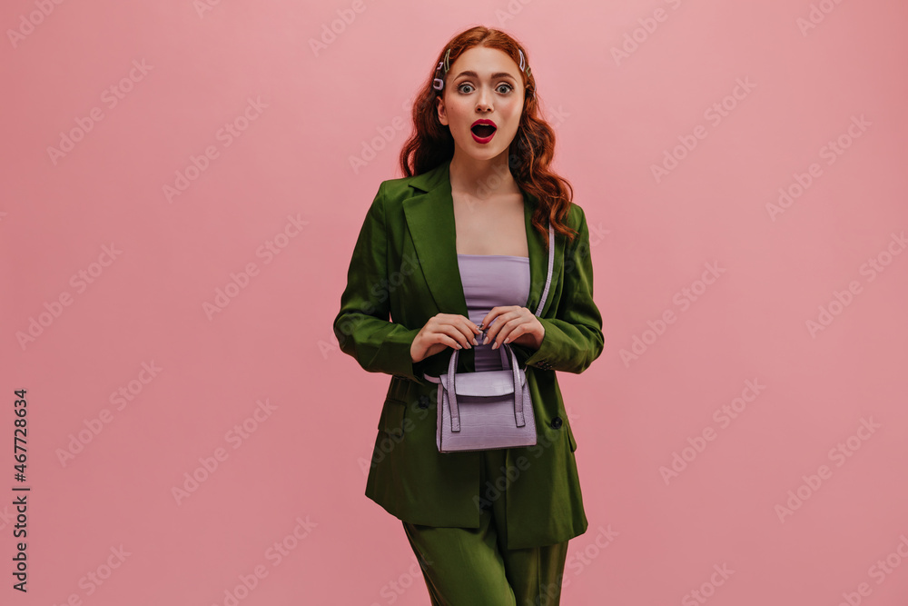 Lovely caucasian young lady with surprised face expression with open mouth over isolated background. Girl with bulging eyes looks at camera holding small handbag in front of her. Lovely caucasian young lady with surprised face expression with open mouth over isolated background. Girl with bulging eyes looks at camera holding small handbag in front of her.