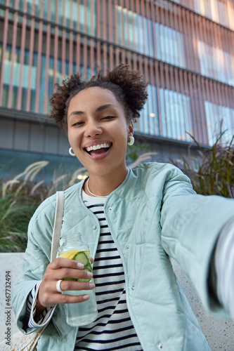 Happy optimistic girl with two hair buns dressed in jacket enjoys free time and walking in city holds bottle of detox drink makes selfie poses against modern building has fun during daytime. photo