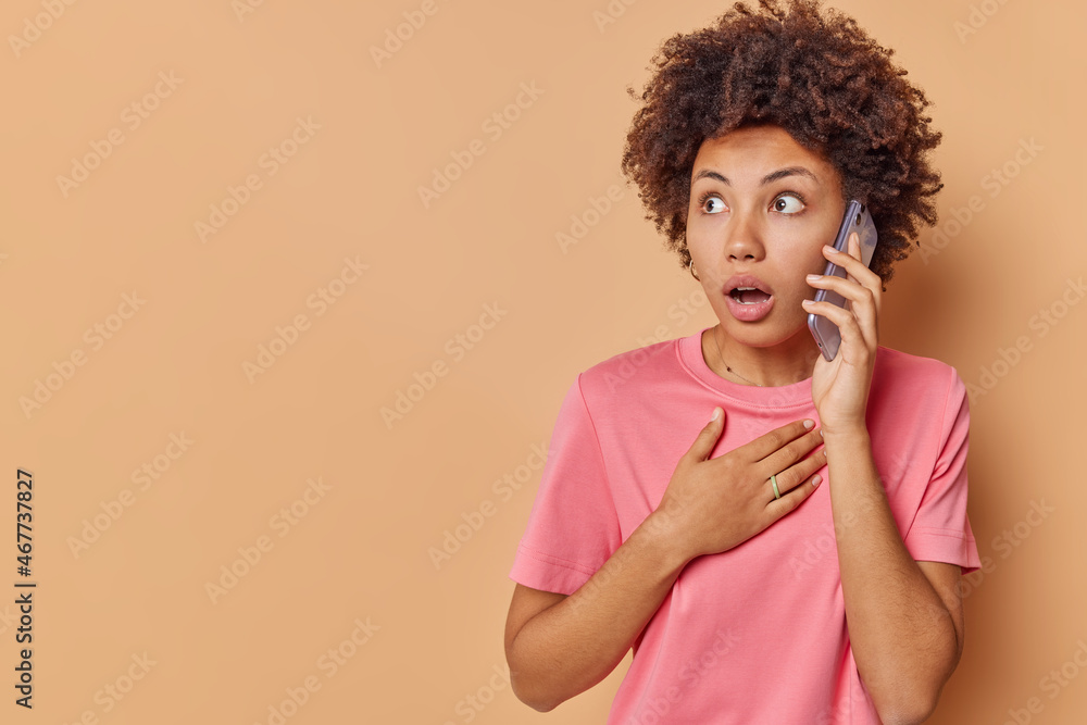 Shocked terrified woman with curly hair talks via phone listens information hears unbelievable news with surprised expression keeps hand on chest concentrated away poses against beige background Shocked terrified woman with curly hair talks via phone listens information hears unbelievable news with surprised expression keeps hand on chest concentrated away poses against beige background
