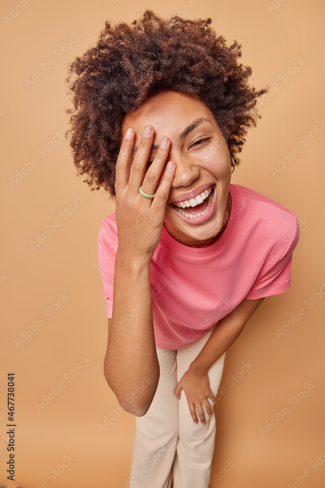 Joyful curly haired young woman laughs sincerely keeps hand on face stands indoor against beige background dressed in casual clothes poses against brown background expresses positive emotions. Joyful curly haired young woman laughs sincerely keeps hand on face stands indoor against beige background dressed in casual clothes poses against brown background expresses positive emotions.