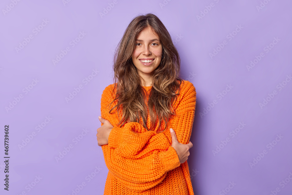 Positive tender young European woman with dark hair embraces herself wears warm knitted orange sweater smiles positively poses against purple background feels chilly walking during cold weather Positive tender young European woman with dark hair embraces herself wears warm knitted orange sweater smiles positively poses against purple background feels chilly walking during cold weather