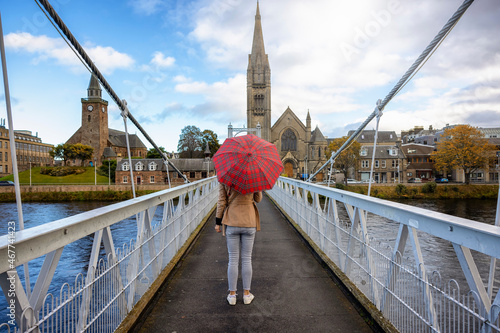A tourist woman with a scotish pattern umbrella walks over the Greig Street Footbridge towards the city of Inverness, Scotland photo