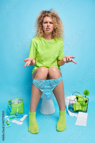 Hesitant clueless woman raises palms in bewilderment poses on toilet bowl suffers from diarrhea wears t shirt panties and socks poses at lavatory room against blue background. Human necessities