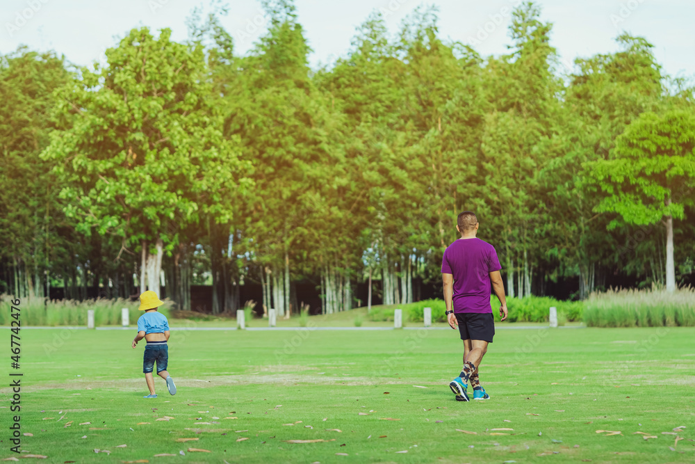 Back view of Asian father and son walking on lawn through green garden. Dad and son walking together in park. Happy family spending time together outside in green nature. Enjoying nature outdoors.