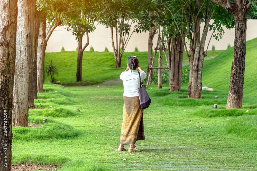 Asian female walking and use smartphone to take a selfie on pathway of park with tall green trees. A woman uses cell phone to take pictures while relaxing in the park. Happy people in green nature. Asian female walking and use smartphone to take a selfie on pathway of park with tall green trees. A woman uses cell phone to take pictures while relaxing in the park. Happy people in green nature.