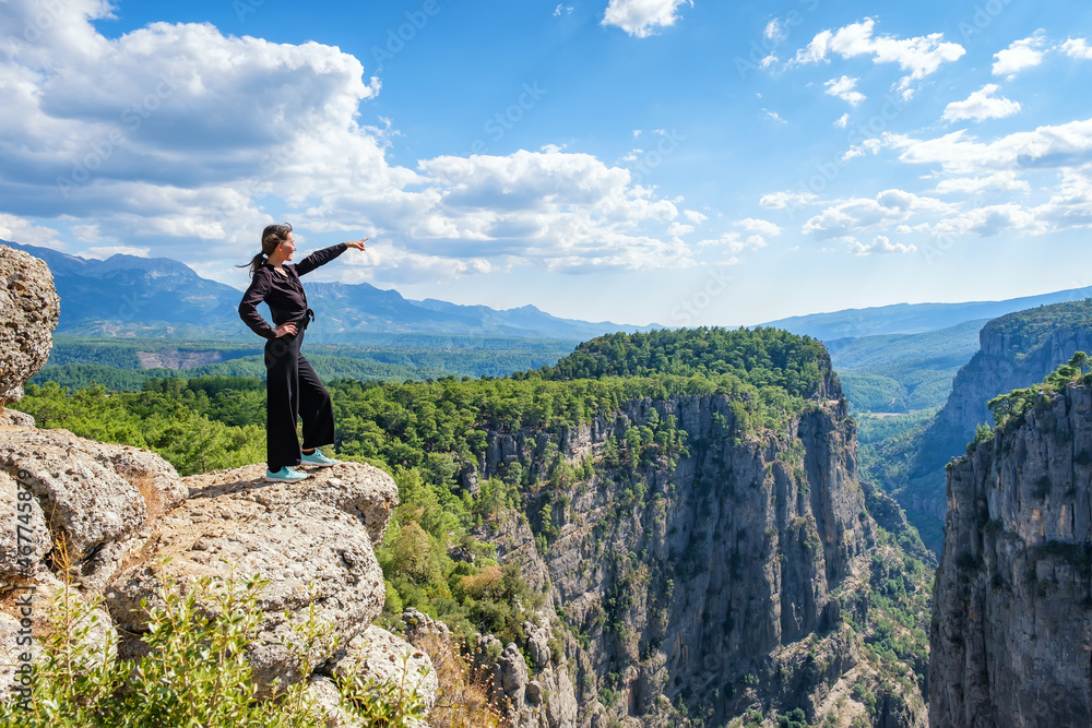Obraz premium Side view of smiling female standing on edge of rocky cliff in highlands and pointing away on summer day