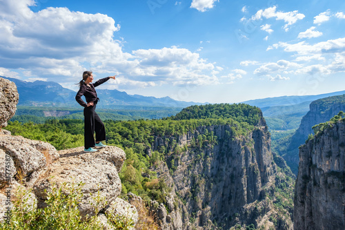 Side view of smiling female standing on edge of rocky cliff in highlands and pointing away on summer day