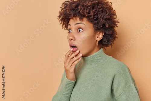 Startled curly haired woman holds chin and stares bugged eyes away holds breath from amazement notices something terrible wears casual jumper isolated over beige background blank copy space.