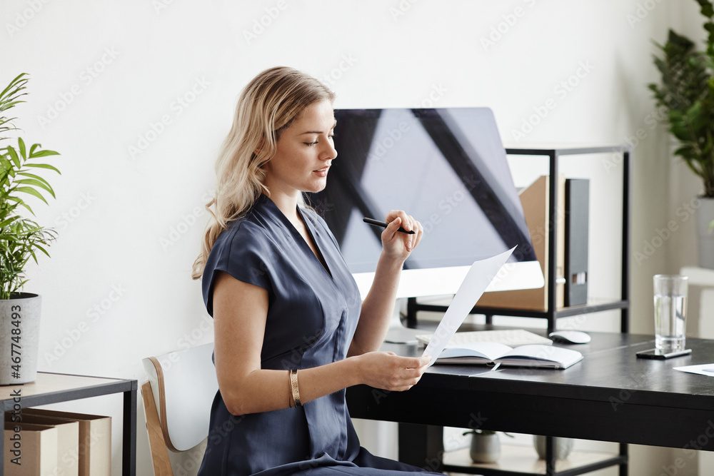 Side view portrait of elegant blonde businesswoman reading documents while working at desk in office, copy space Side view portrait of elegant blonde businesswoman reading documents while working at desk in office, copy space
