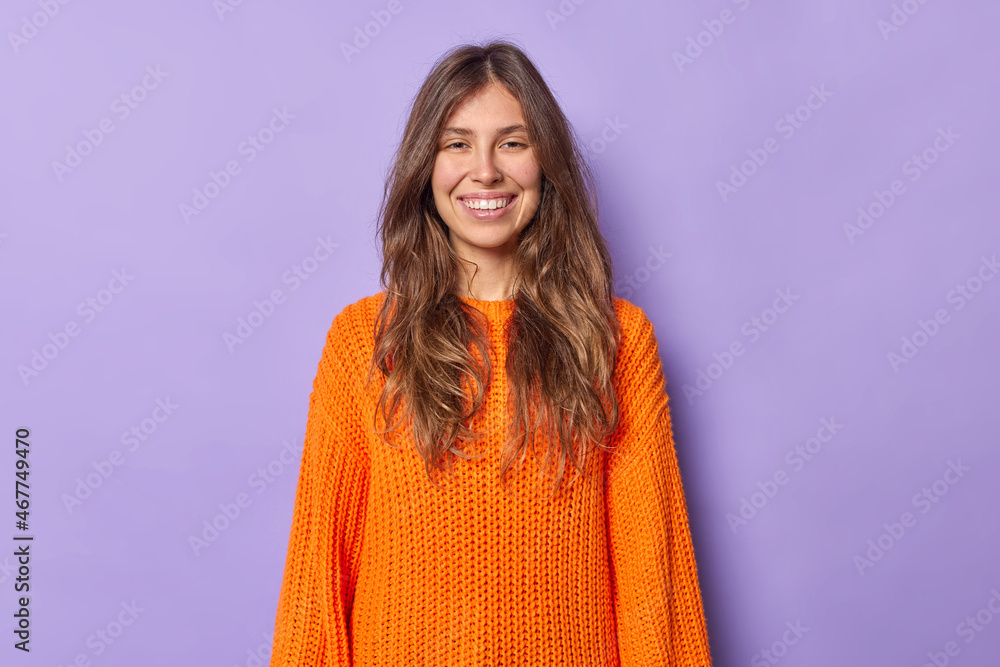 Fototapeta premium Portrait of pretty young European woman with long dark hair smiles pleasantly wears casual orange knitted sweater isolated over purple background shows white teeth. People and emotions concept