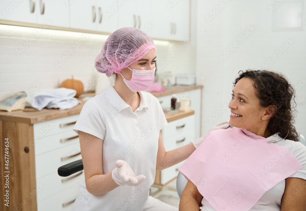 Fototapeta premium Dentist hygienist, dental assistant wearing pink medical mask and surgical gloves preparing woman patient to medical check-up, giving consultation about oral hygiene in dentistry modern clinic