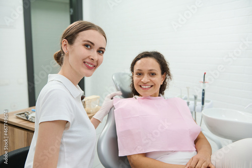 Attractive confident woman  dentist hygienist cutely smiles looking at camera beside female patient sitting in dentists chair before receiving dental treatment in dentistry clinic. Oral hygiene care