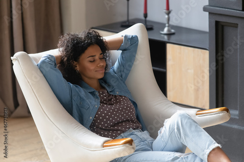 Peaceful smiling young Black woman resting in cozy soft armchair, breathing fresh air, enjoying being at comfortable home. Millennial girl reloading mind, practicing stress relief, mindfulness