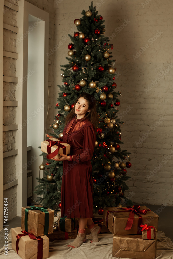 Portrait of a girl in a beautiful burgundy dress near a new year tree with gifts
