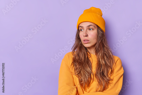 Horizontal shot of pensive long haired European woman concentrated away being deep in thoughts considers something wears orange hat and jumper poses agaist purple background with blank space