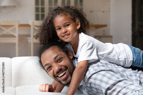 Cheerful African American daughter girl resting on happy dads back, looking at camera, smiling. Portrait of daddy lying on sofa at home, piggybacking child, enjoying leisure time, playing active games © fizkes