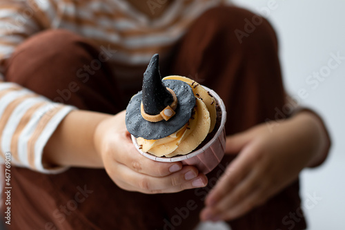 A little boy sits on the windowsill and eats a cupcake in the form of a witch's hat. Halloween concept
