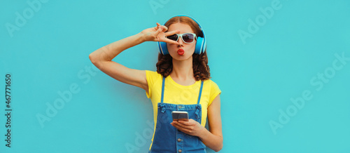 Fashionable portrait of stylish young woman in headphones listening to music with smartphone on blue background