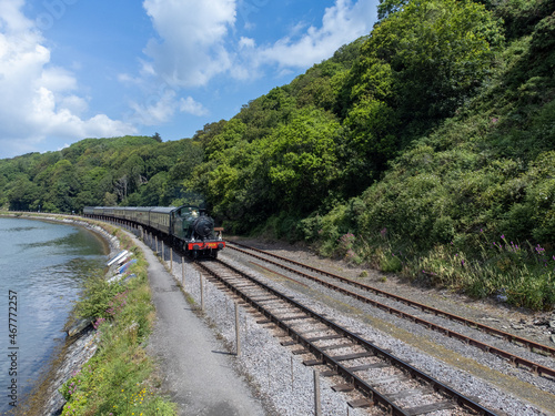 Photos steam train on train track next to water and near a hill with trees and blue sky