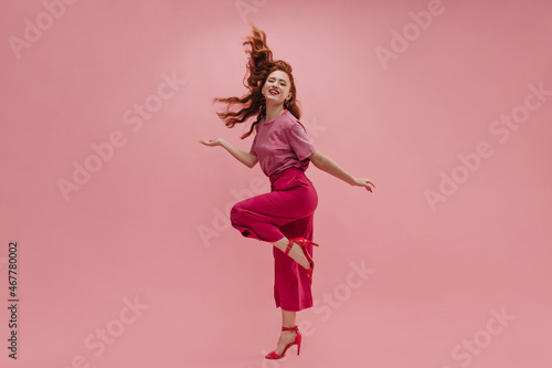 In full growth, fantastic caucasian young girl stands sideways with leg curled up at knee on pink studio background. Redhead model waving long hair smiling at camera, wearing t-shirt, pants and heels.