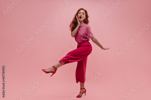 Attractive young woman raising leg in front of in surprise looks at camera in studio shot. Model with red wavy hair covers open mouth with hand, wearing T-shirt, pants. Concept admiration for preppy