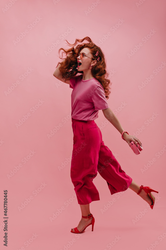 Full-length portrait of cheerful caucasian young lady with flying gait walking on photo indoors. Her red wavy hair blows in wind. Beauty with open mouth in glasses, T-shirt, pants and sandals.