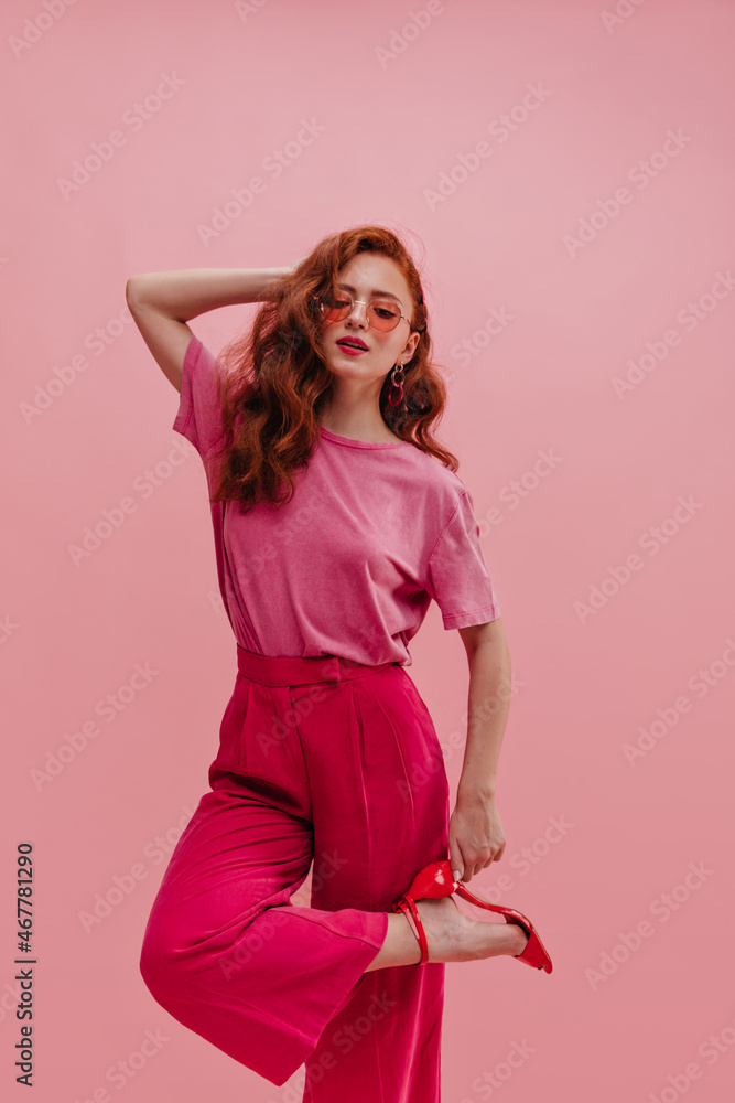 Not full-length caucasian young red-haired girl posing holding back of her head in closed space. Beauty is holding on to heel of red shoes, wearing T-shirt tucked into pants and translucent glasses. Not full-length caucasian young red-haired girl posing holding back of her head in closed space. Beauty is holding on to heel of red shoes, wearing T-shirt tucked into pants and translucent glasses.