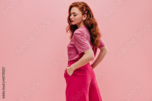 Fashionable caucasian young woman stands half sideways looking down on pink background. Red-haired curly-haired lady with her hands in pants pockets, dressed in casual clothes. Self-confidence concept