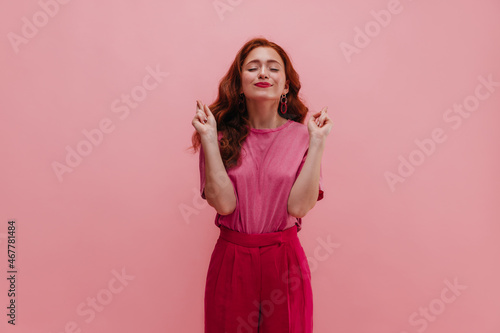 Image of european model with touching smile and closed eyes on pink background. Girl crosses her fingers with both hands in hope of fulfilling wish. Achievement concept.