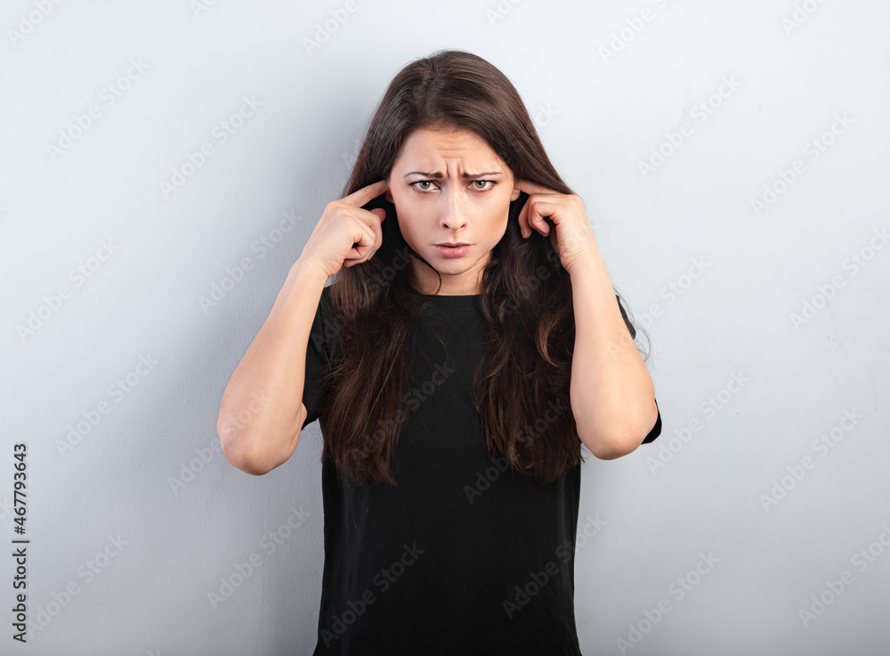 Unhappy stressed grimacing frowns woman not want to listen and covering the ears her fingers on blue background. Unhappy stressed grimacing frowns woman not want to listen and covering the ears her fingers on blue background.