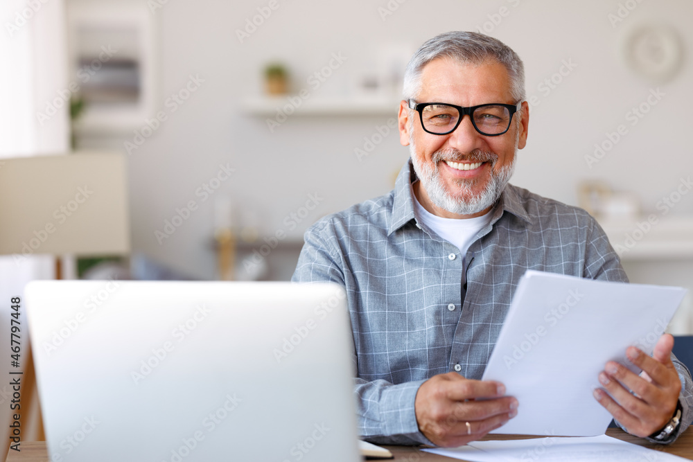 . Handsome smiling senior man reading financial documents while working remotely on laptop at home . Handsome smiling senior man reading financial documents while working remotely on laptop at home