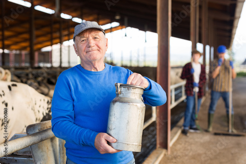 Elderly male owner with milk can standing in stall on background with herd of cows on livestock farm